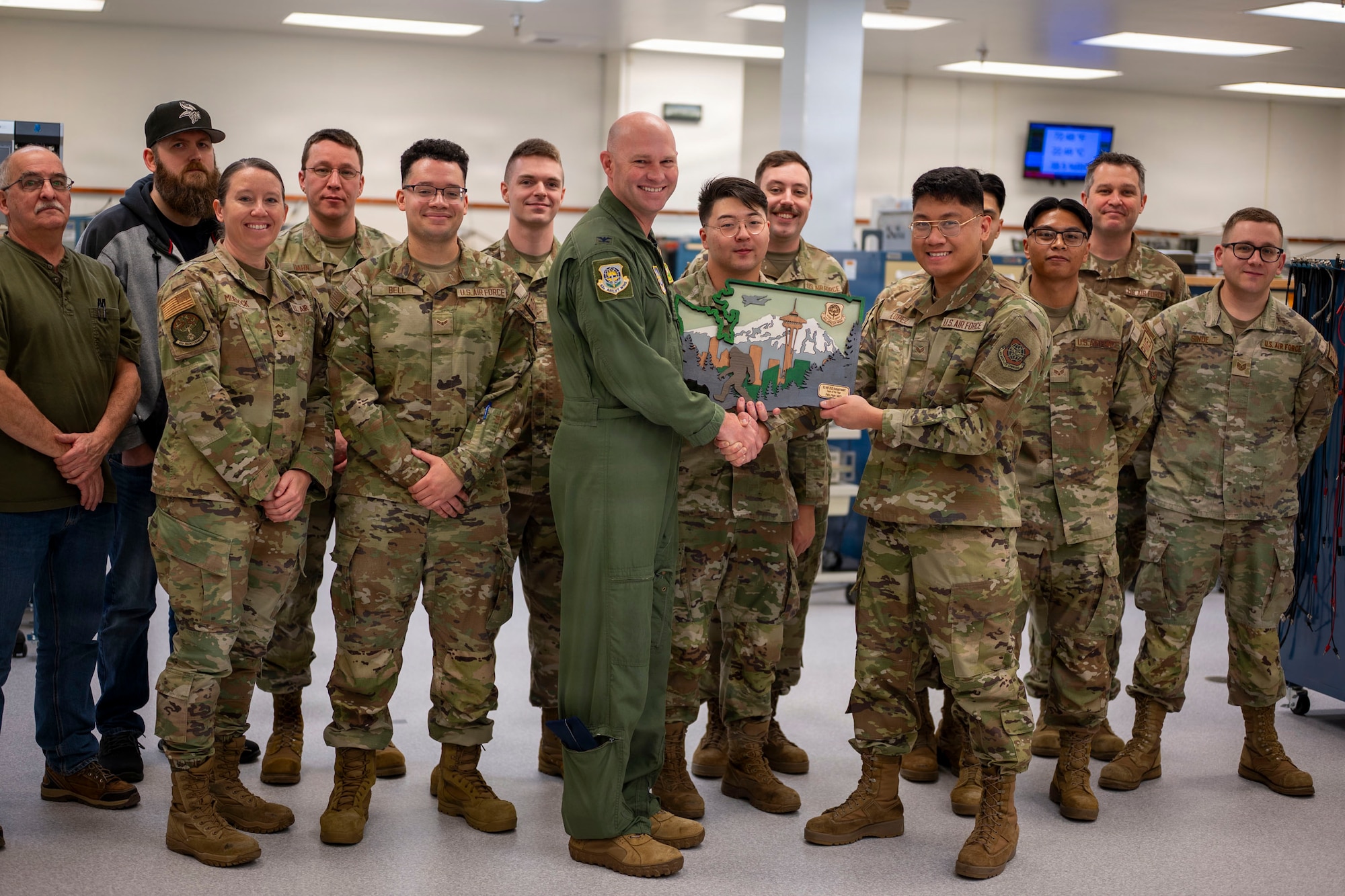 A group of U.S. Air Force Airmen in camouflage face the camera and smile. In the center, an airman in a flight suit hands a plaque to one of the airman in camouflage