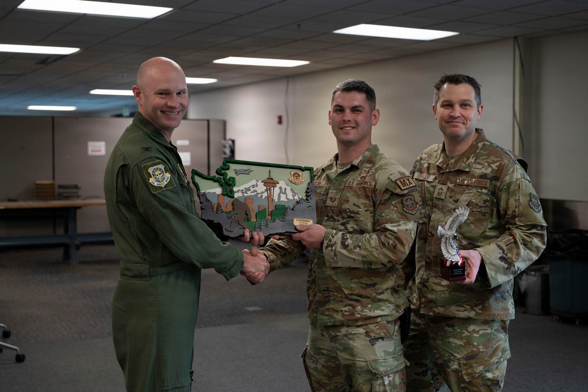 three U.S. Air force airmen face the camera and smile. The airman on the left side hands a plaque to the airman in the center