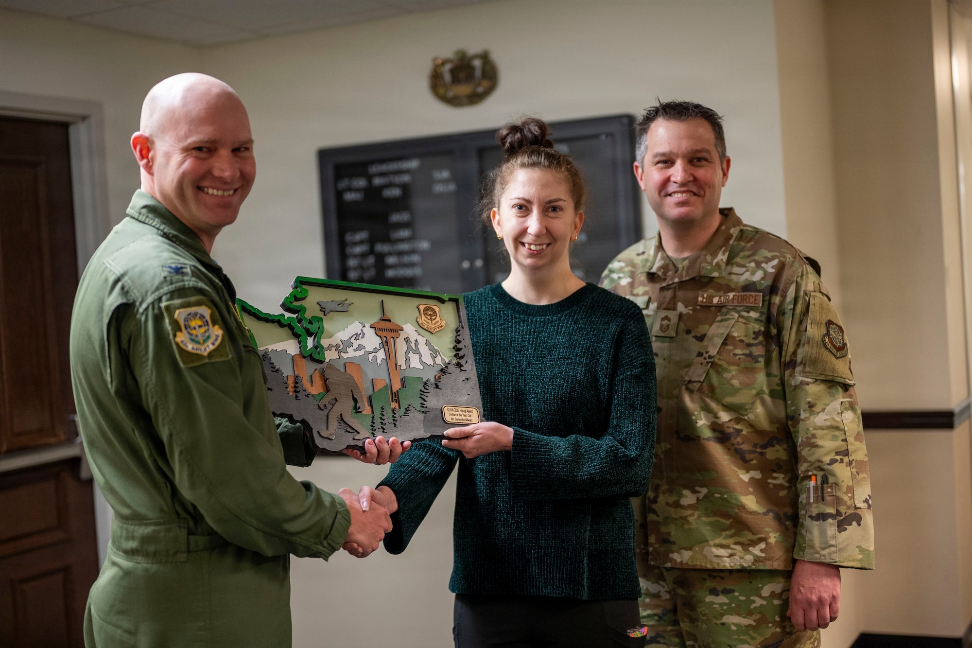 two U.S. Air Force airmen stand on either side of female civilian, all smiling for the camera. The Airmen on the left is handing a plaque to the civilian who is in the center