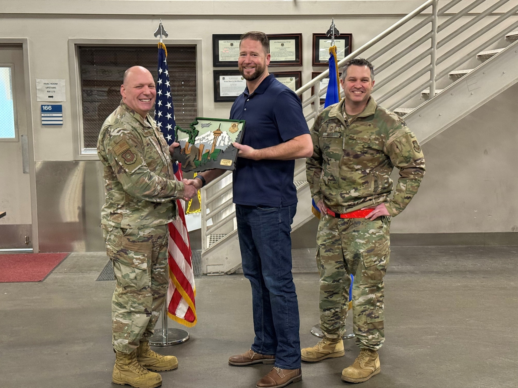 two U.S. Air Force airmen stand on either side of a male in civilian clothes, all smiling for the camera. the airman on the left shakes hands with the civilian in the center while handing him a plaque