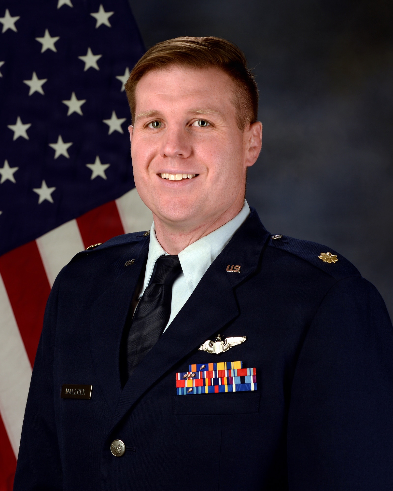 A portrait of a U.S. Air Force male in full service dress with an American flag over his right shoulder. He is smiling.