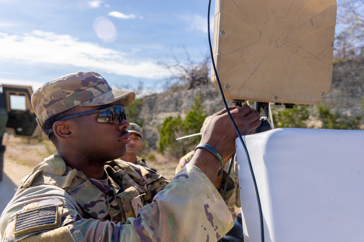 A man wearing a camouflage military uniform works on a piece of military equipment as another man stands nearby and watches him.