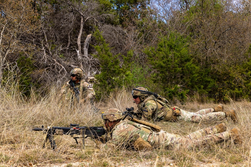 Two men dressed in camouflage military uniforms lay on dry brush covered ground with military automatic weapons in their hands. A third man, also dressed in a camouflage military uniform, is crouching next to a bush and tree.