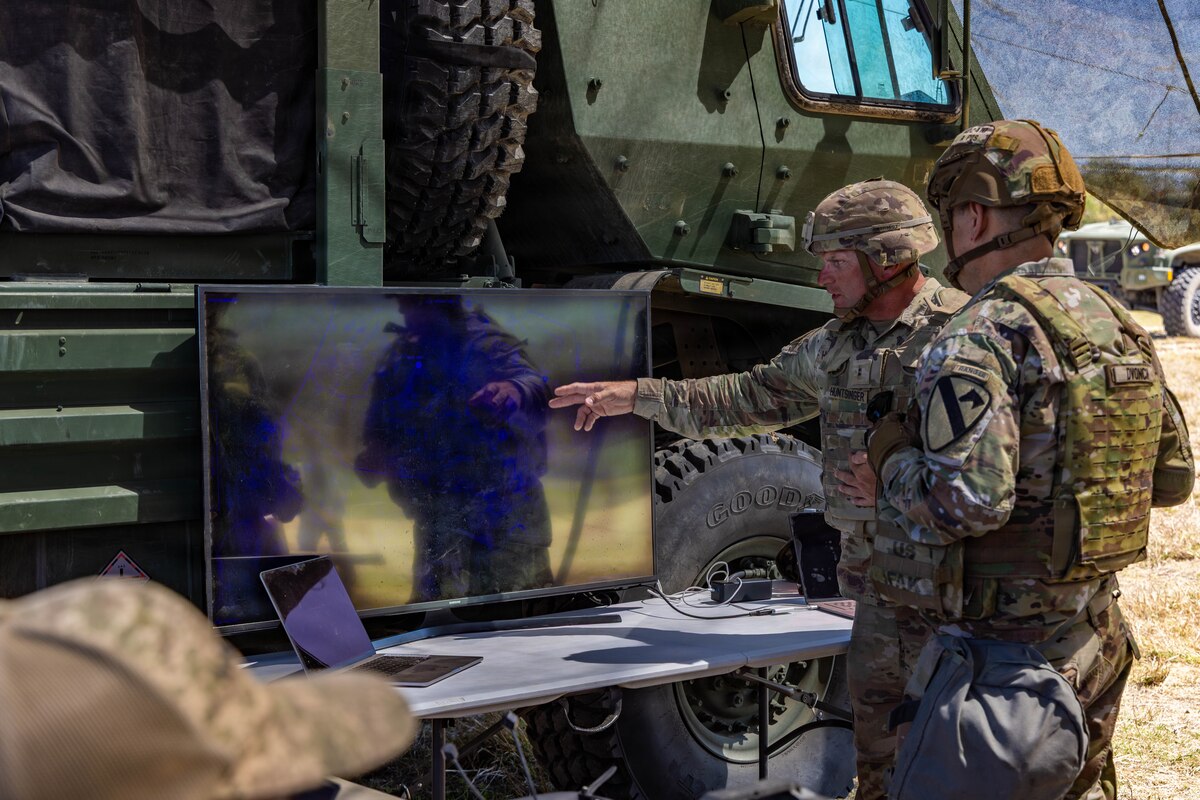 Two men wearing camouflage military uniforms look at a computer screen as one of the men points to something on the screen. There is a large military vehicle behind the computer screen.