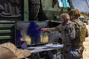 Two men wearing camouflage military uniforms look at a computer screen as one of the men points to something on the screen. There is a large military vehicle behind the computer screen.