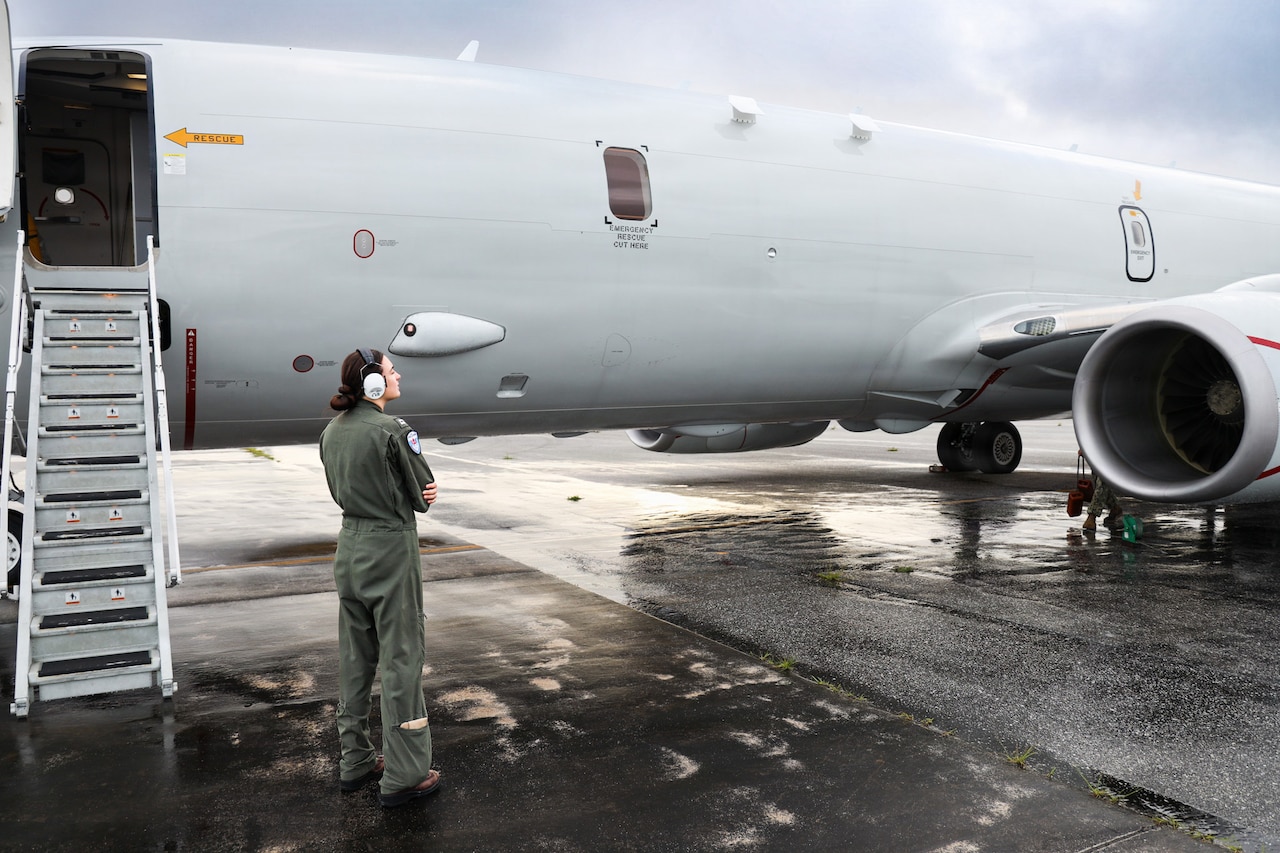 A woman wearing a flight suit looks at a military aircraft sitting on a tarmac with the boarding steps down.