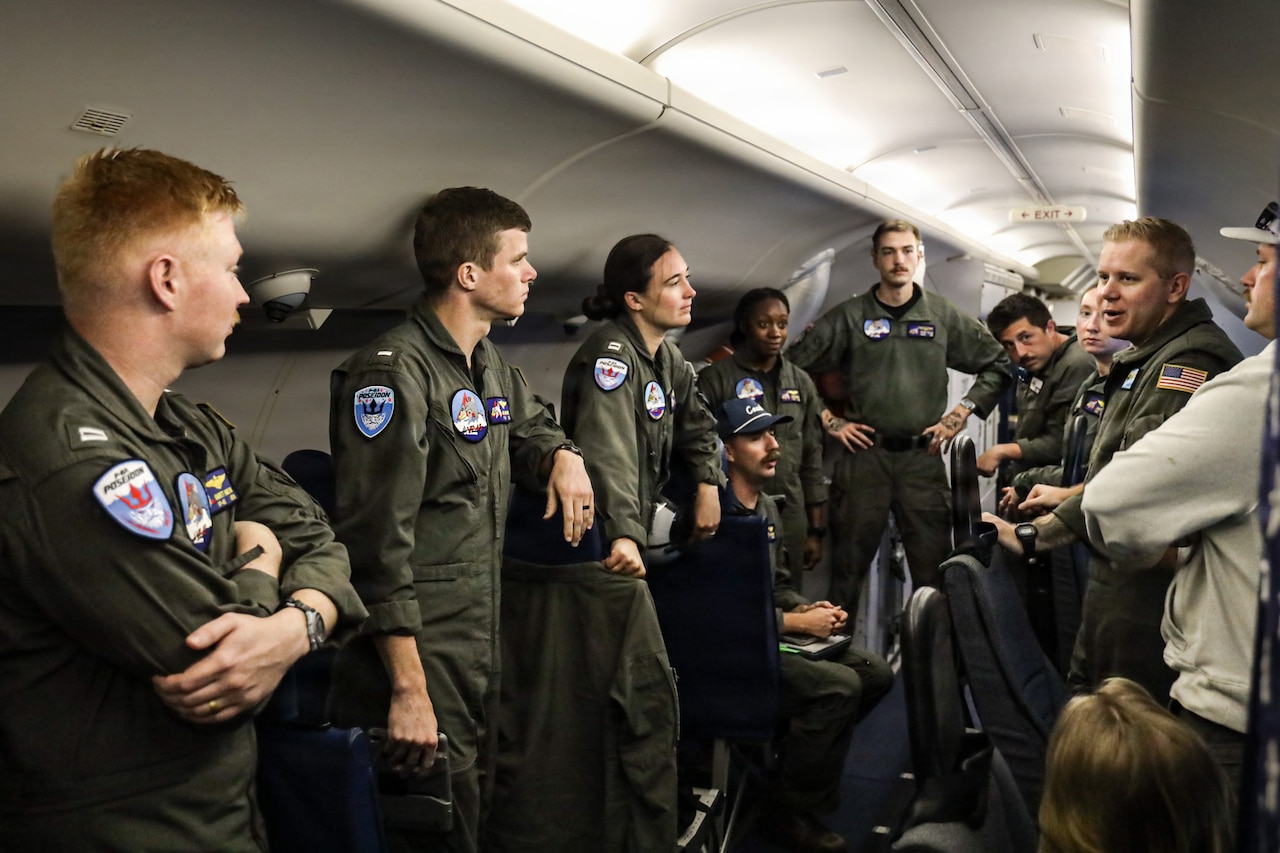 A group of men and women wearing flight suits gather aboard a military aircraft as another person in similar attire speaks to them.