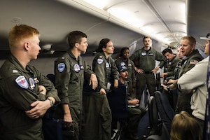A group of men and women wearing flight suits gather aboard a military aircraft as another person in similar attire speaks to them.