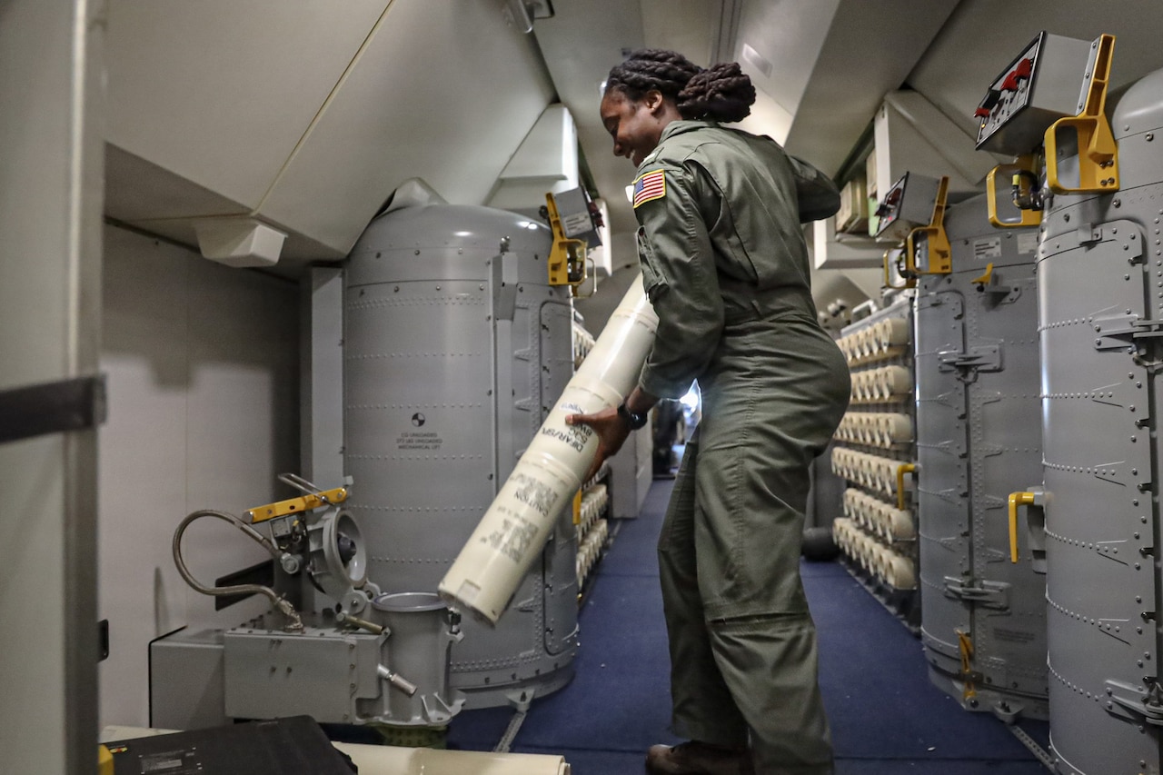 A woman wearing a flight suit moves a submarine tracking buoy into a launcher aboard a military aircraft.