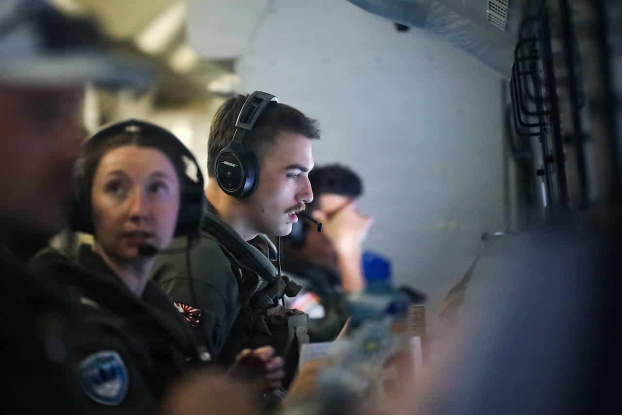 A man wearing a flight suit and headset checks a computer screen while aboard a military aircraft. There are three other people in similar attire sitting around him working at their stations.