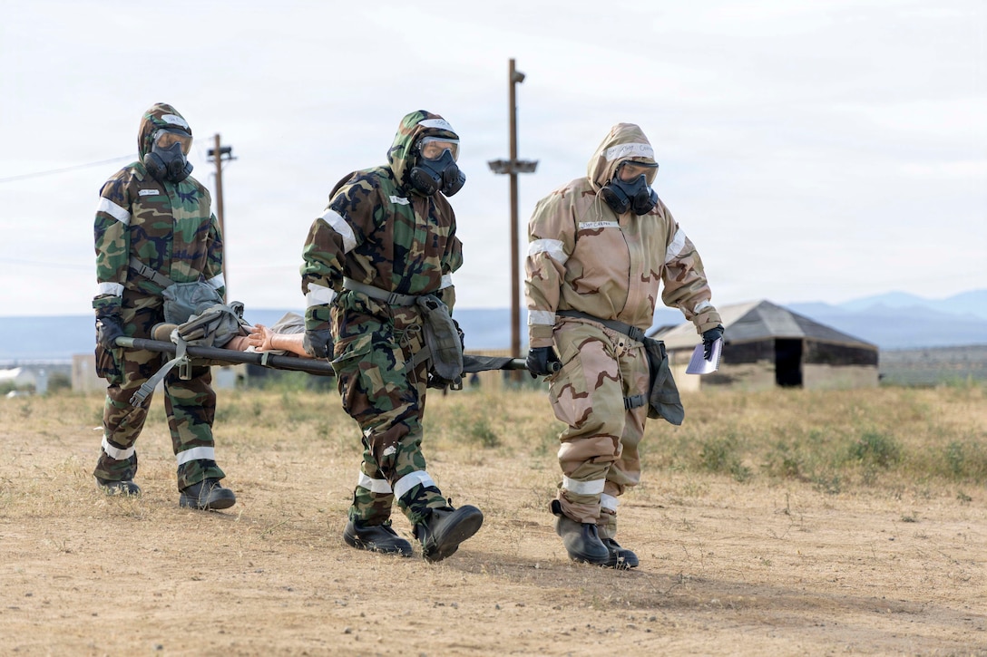 Three airmen in gas masks and protective camouflage suits carry a dummy on a military stretcher in a field with mountains in the background.