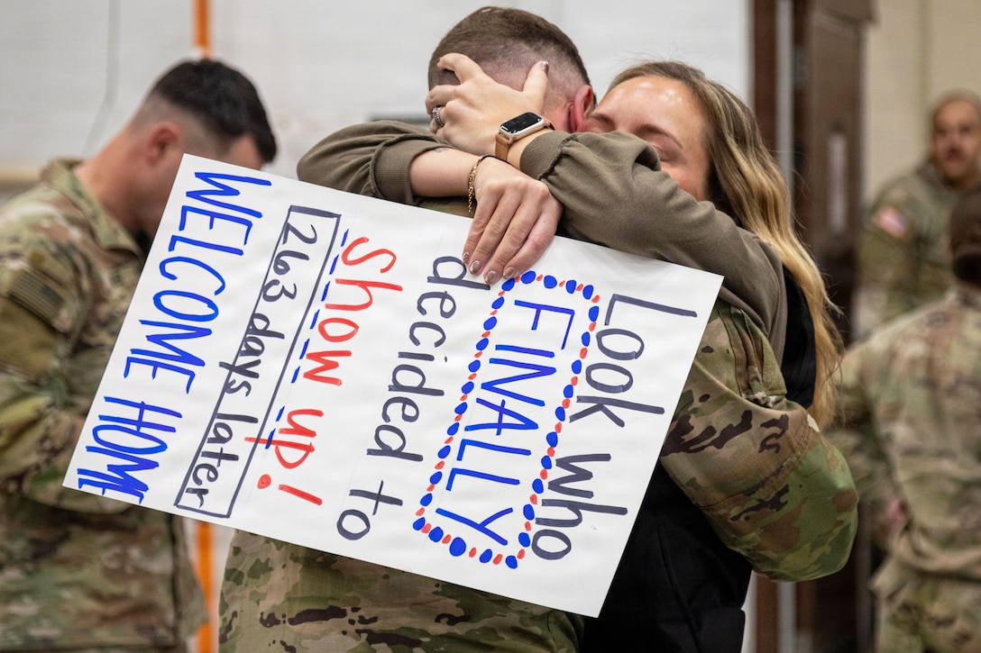 A loved one holding a homemade welcome sign hugs a soldier as fellow soldiers gather in the background.