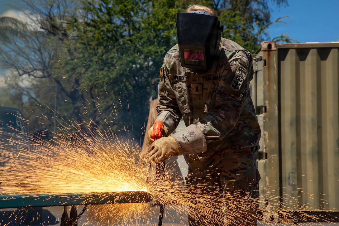 Sparks fly as a soldier in protective gear cuts through metal with a torch on a table during the day.