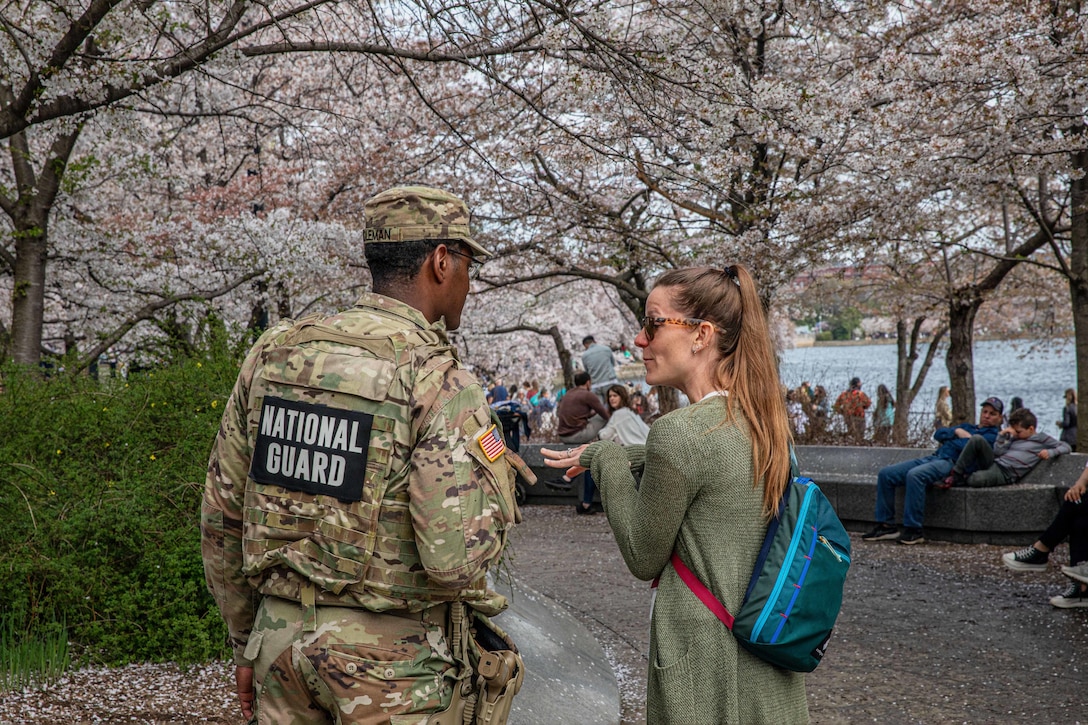 A soldier speaks to a tourist as fellow tourists sit under cherry blossoms near a body of water.