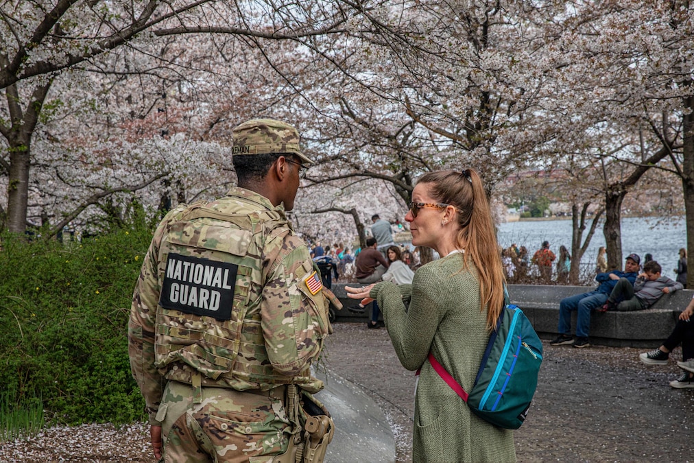 A soldier speaks to a tourist as fellow tourists sit under cherry blossoms near a body of water.
