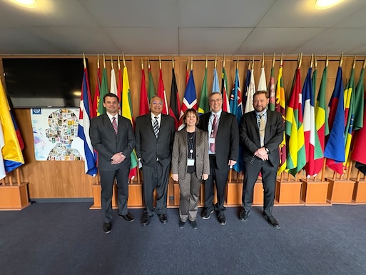 Five people stand inside in front of flags posing for a photo.