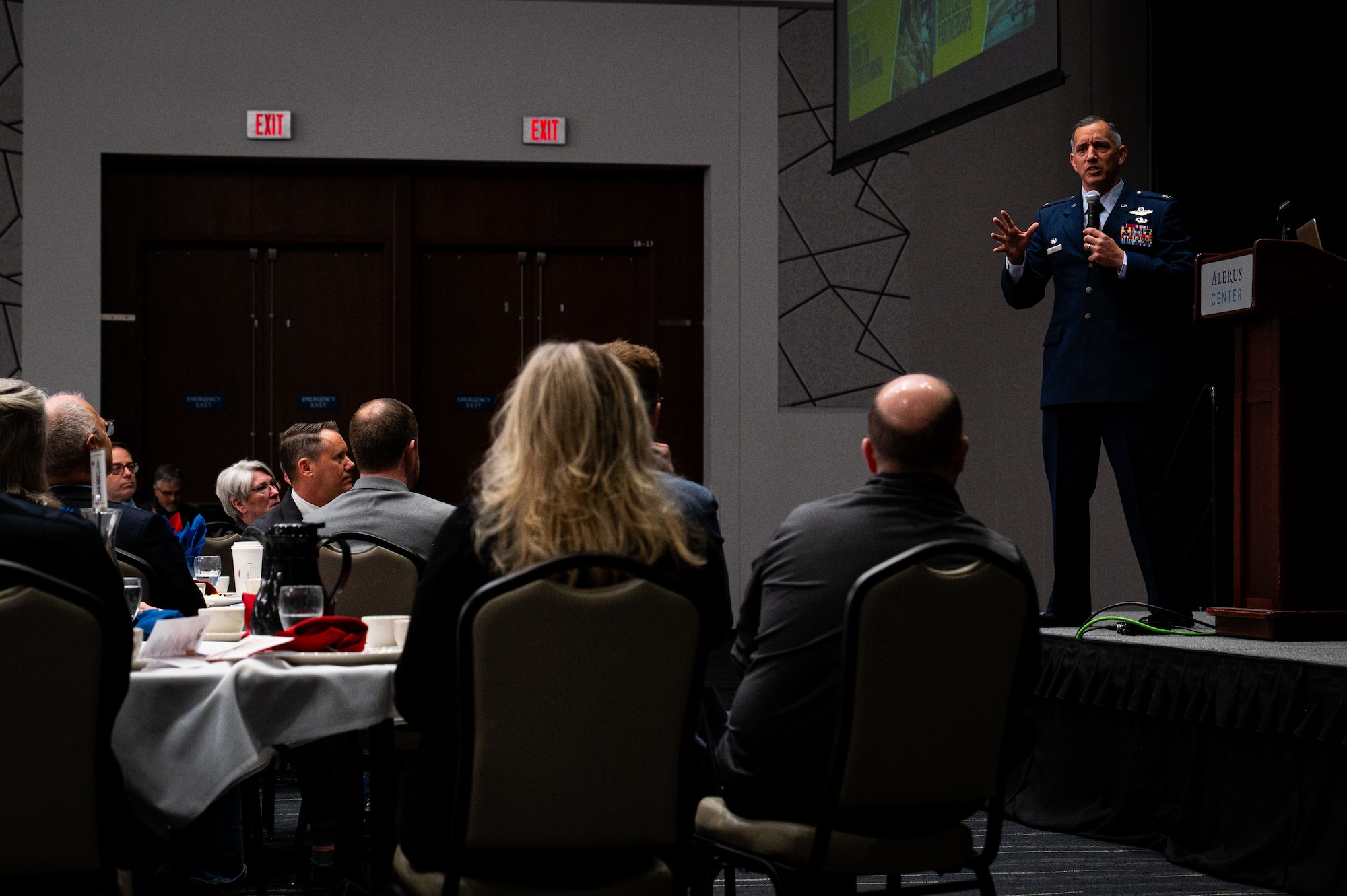 A photo of a man standing on a stage speaking into a microphone in front of a crowd.