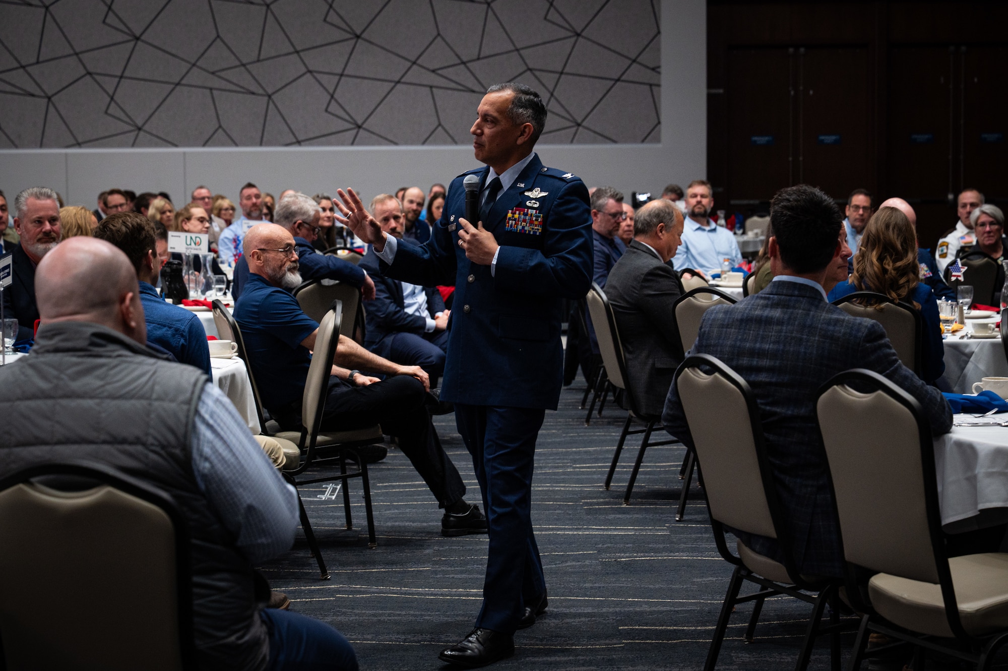 A photo of a man standing with a crowd of people sitting at table around him, while he speaks into a microphone.