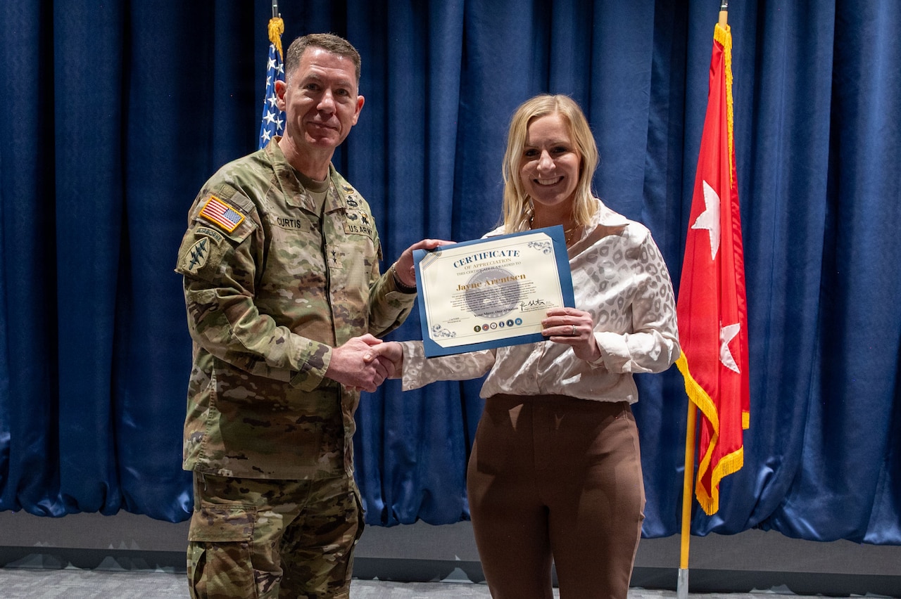 A man in a camouflage military uniform shakes hands and presents a certificate to a woman in business attire on a stage. Behind them are a blue curtain, an American flag and a red flag with two white stars in the center.