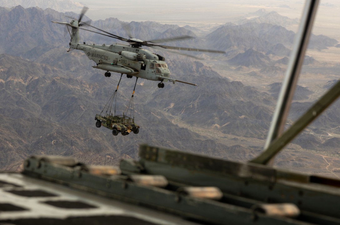 A helicopter lifts two military vehicles that are suspended from it by cables while flying above mountains.