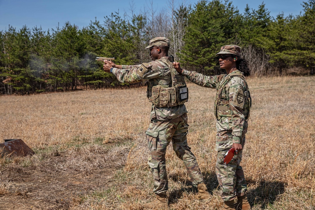 Two soldiers stand outside on grass, with trees in the background. One soldier aims a pistol and the other holds a device in the air behind him.