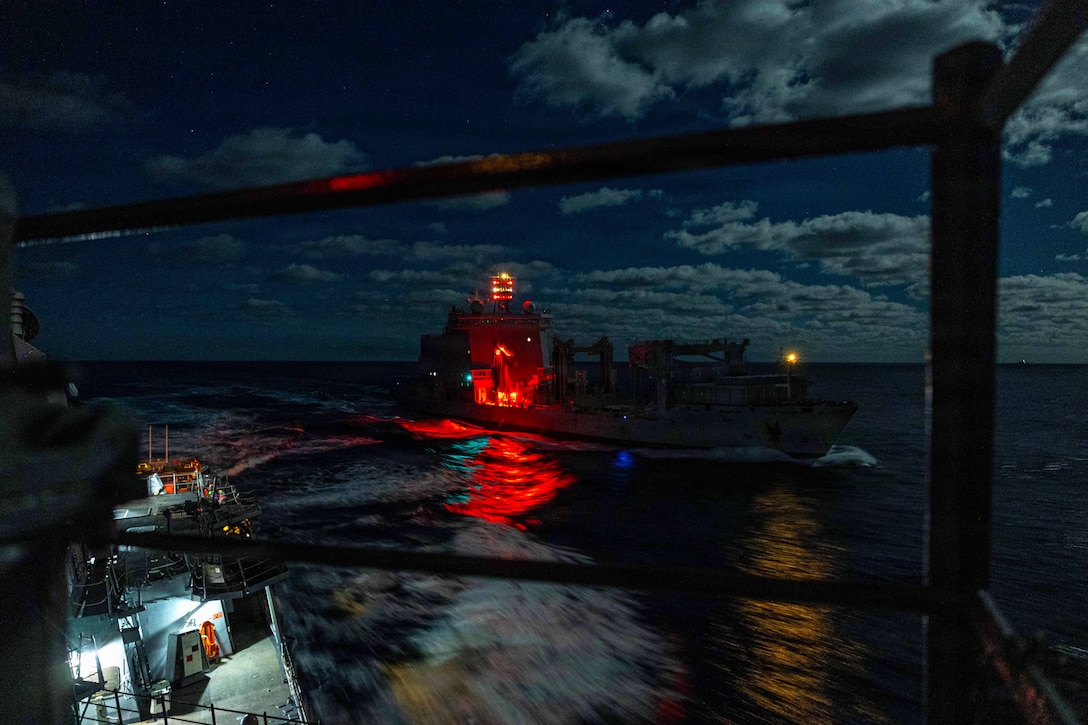 One ship moves away from another ship at sea, and lights from the ships are reflected on the water.