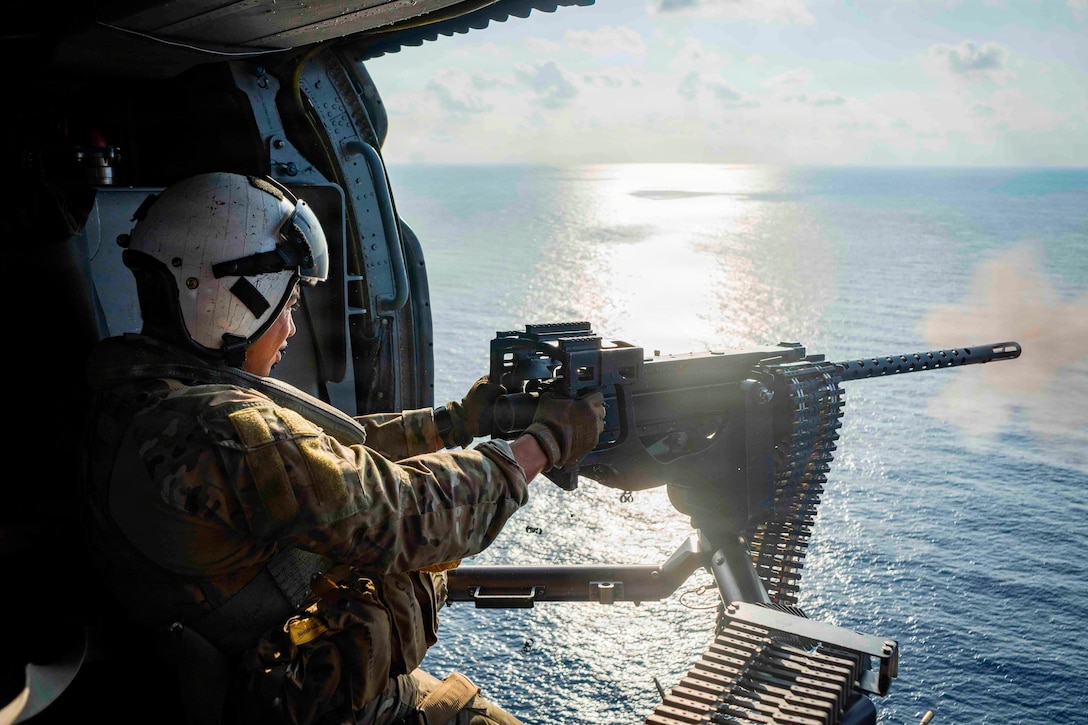 A sailor wearing a helmet and a camouflage uniform fires a machine gun from a helicopter above the sea.