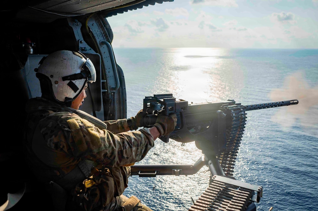 A sailor wearing a helmet and a camouflage uniform fires a machine gun from a helicopter above the sea.