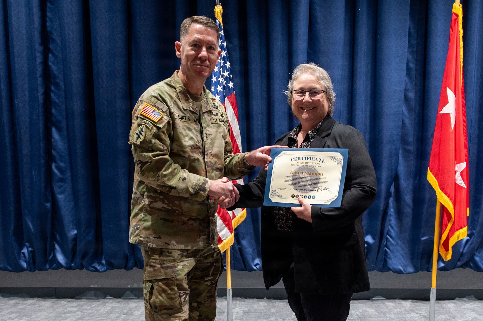 A man in a camouflage military uniform shakes hands and presents a certificate to a woman in business attire on a stage. Behind them are a blue curtain, an American flag and a red flag with two white stars in the center.