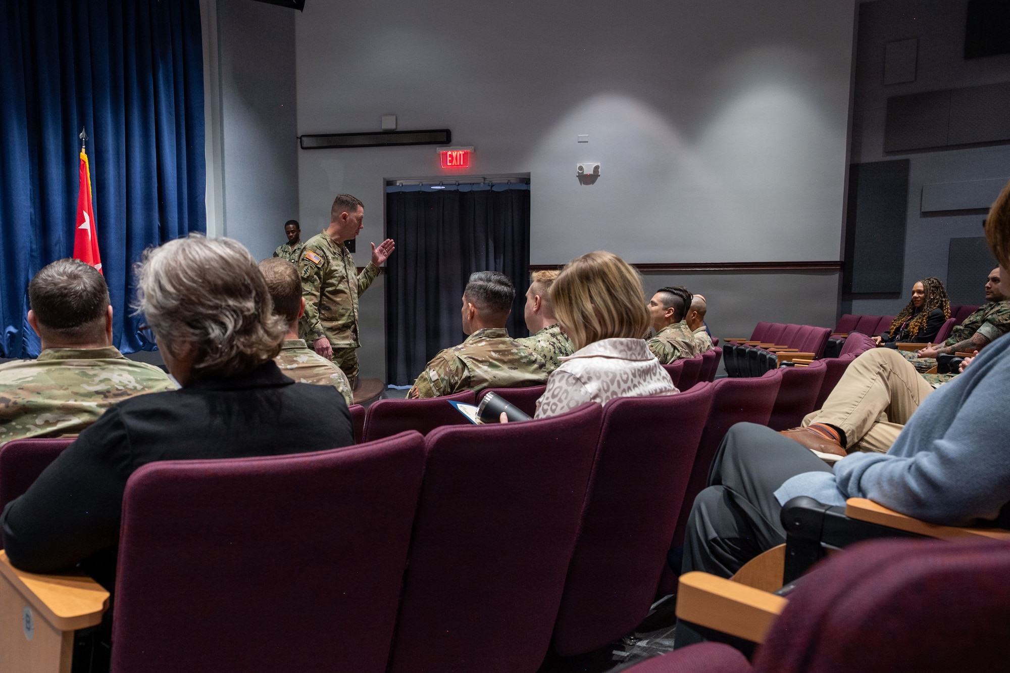 A man in a camouflage military uniform gestures with his hands and speaks to an audience inside an auditorium.