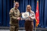 A man in a camouflage military uniform shakes hands and presents a certificate to a woman in business attire on a stage. Behind them are a blue curtain, an American flag and a red flag with two white stars in the center.