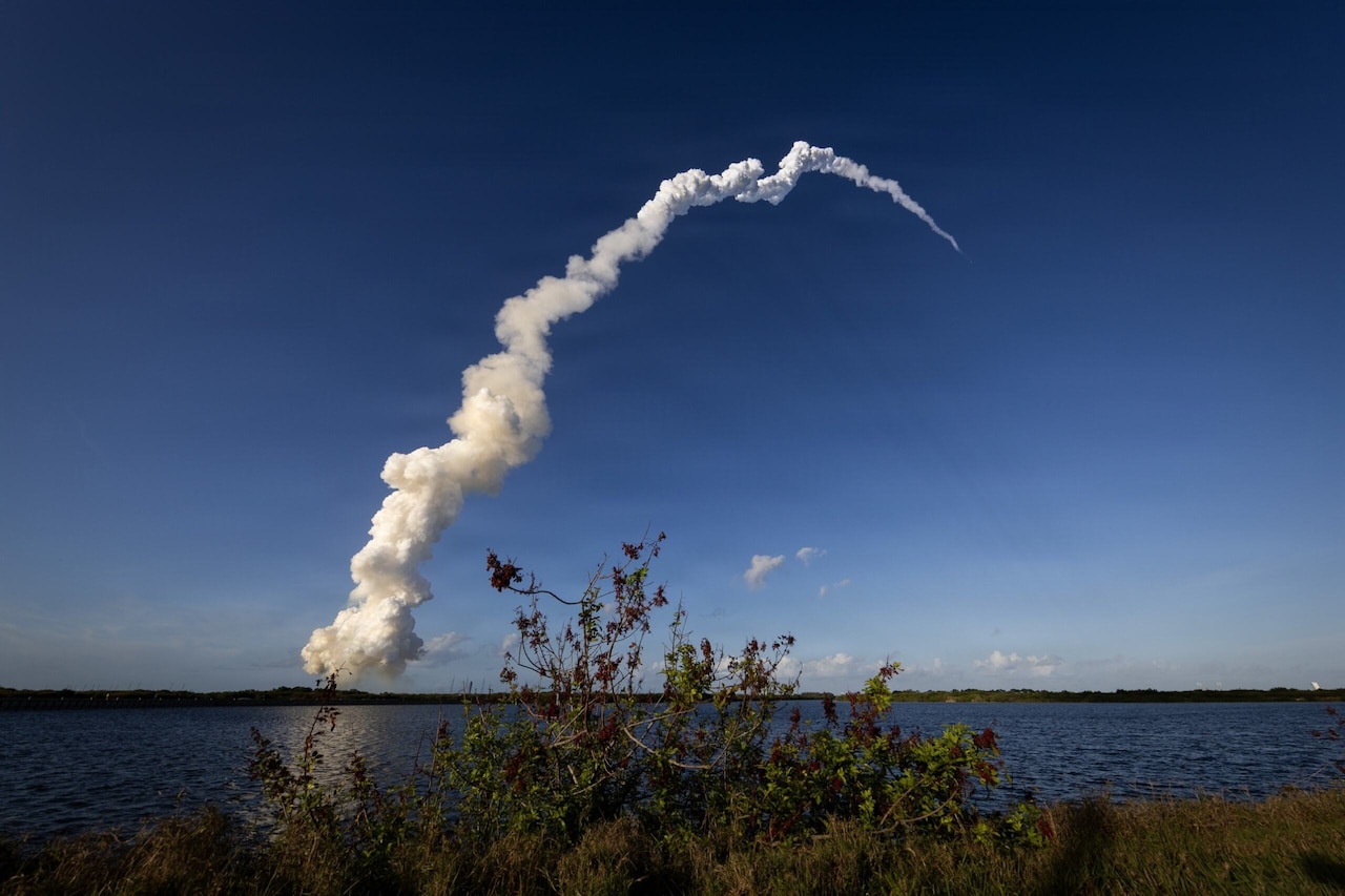 A rocket flies through the sky as it leaves a long trail of smoke behind it, leading back to the launch pad. There are shrubs and a large body of water in the foreground.