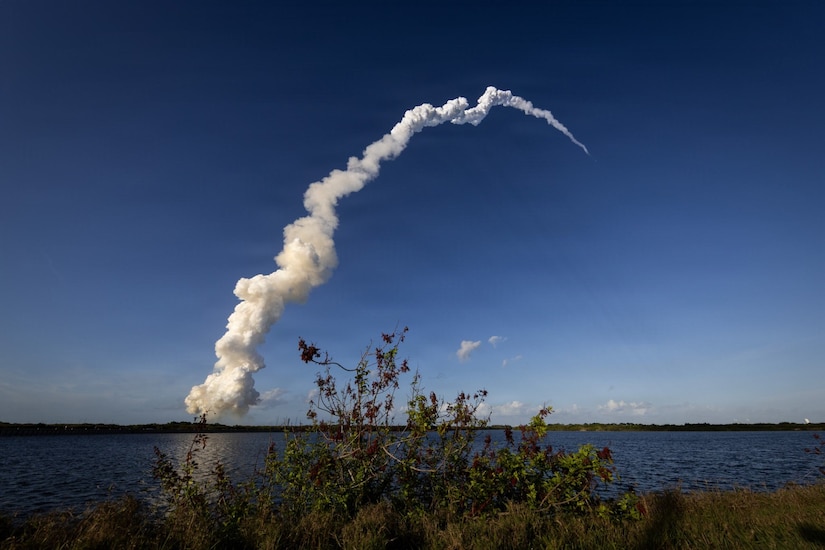A rocket flies through the sky as it leaves a long trail of smoke behind it, leading back to the launch pad. There are shrubs and a large body of water in the foreground.