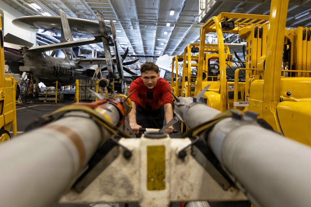 A sailor in a red shirt pulls ordnance in a hangar bay in between parked equipment.