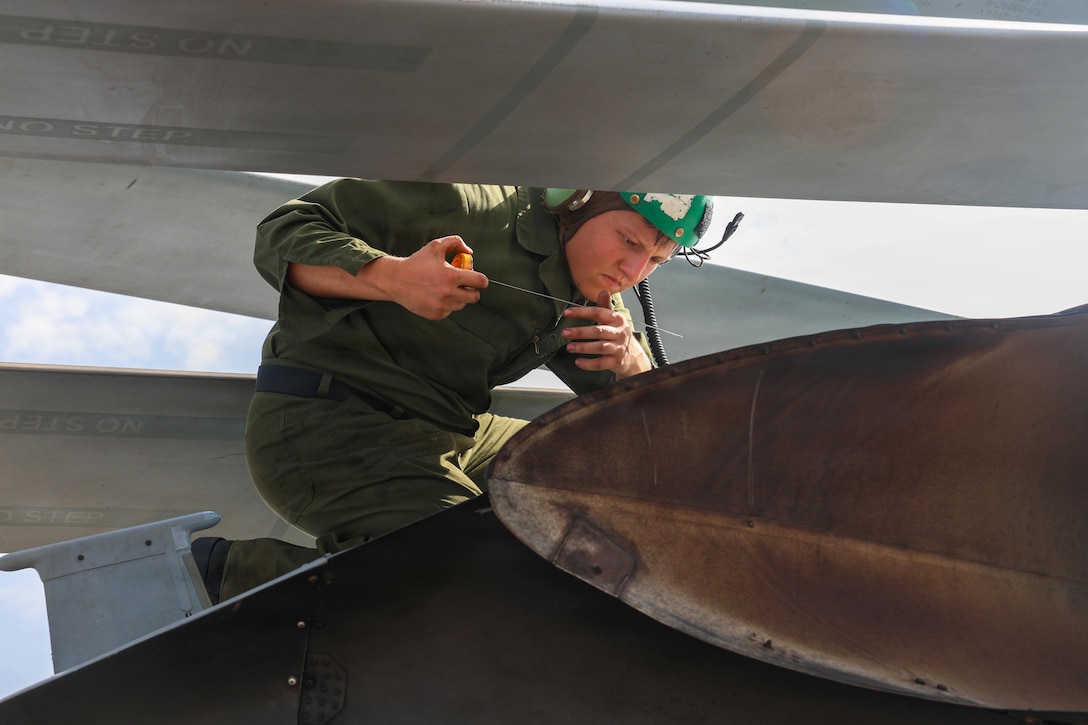 A sailor wearing a green helmet kneels on a helicopter while checking its oil during the day.