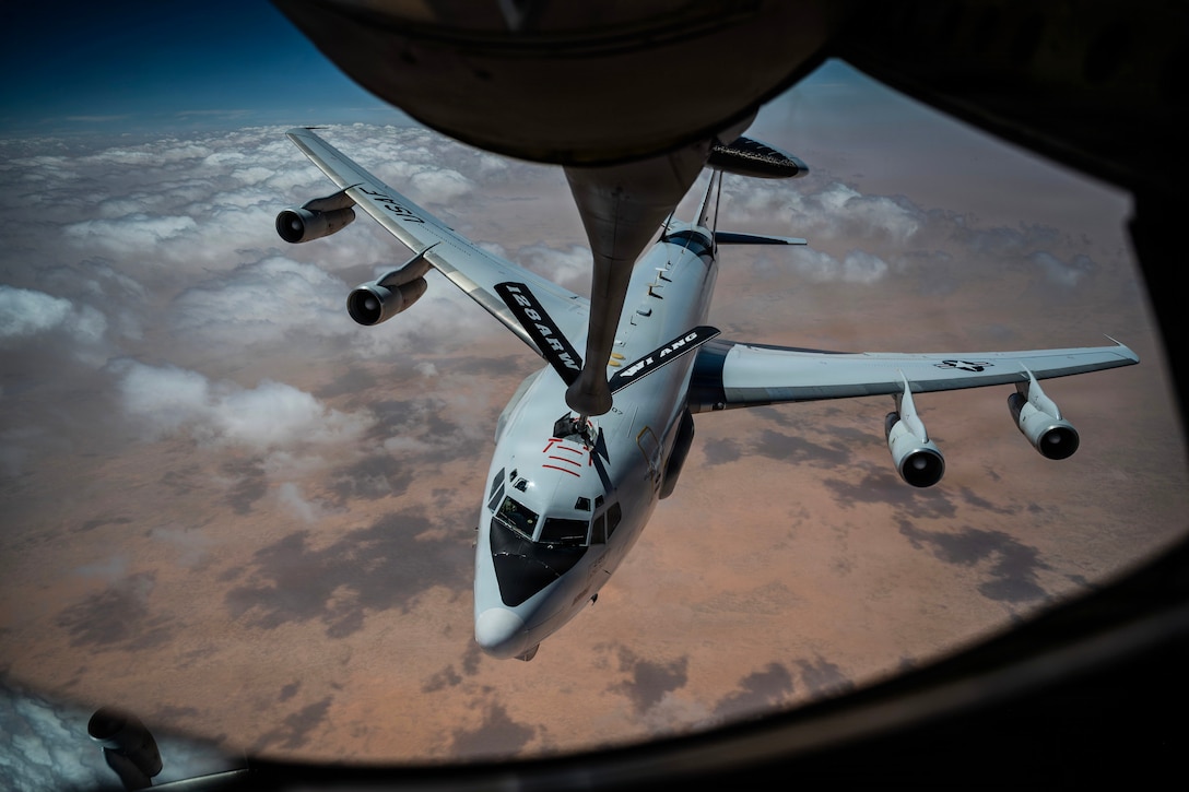 A partially visible aircraft extends its boom to another aircraft as they fly through the clouds over a desert-like area.