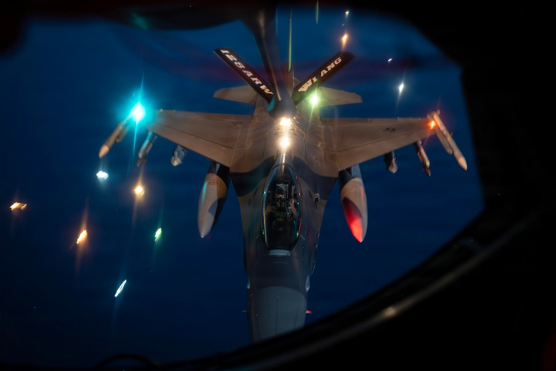 An aircraft extends its boom to another aircraft in a dark sky illuminated by red, green and white lights.