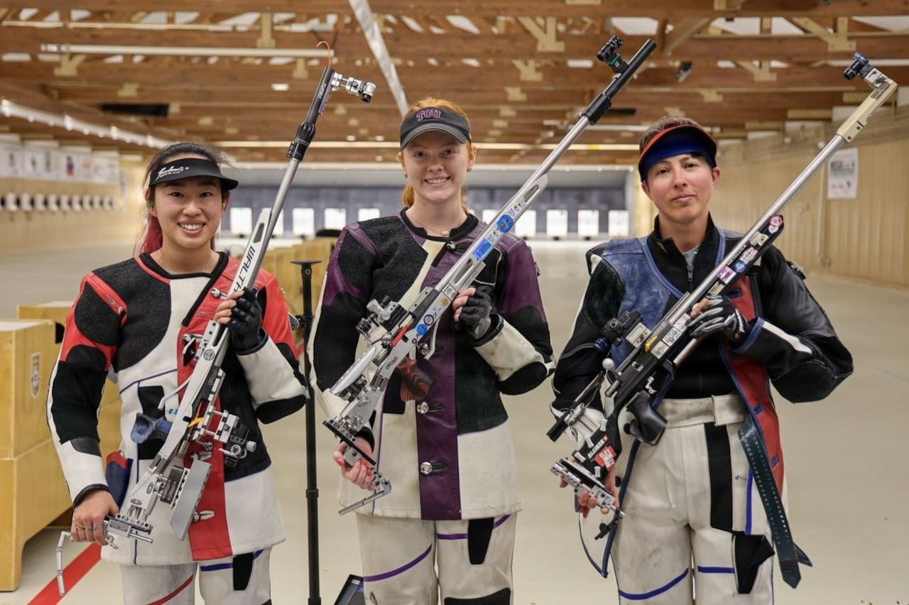 Three women dressed in competitive rifle shooting gear hold rifles up at an angle while posing for a photo.