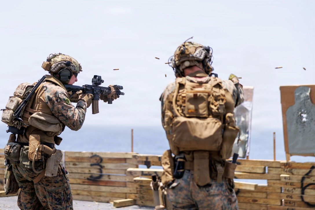 Ammo flies in the air as Marines in tactical gear fire weapons at targets aboard a ship during the day.