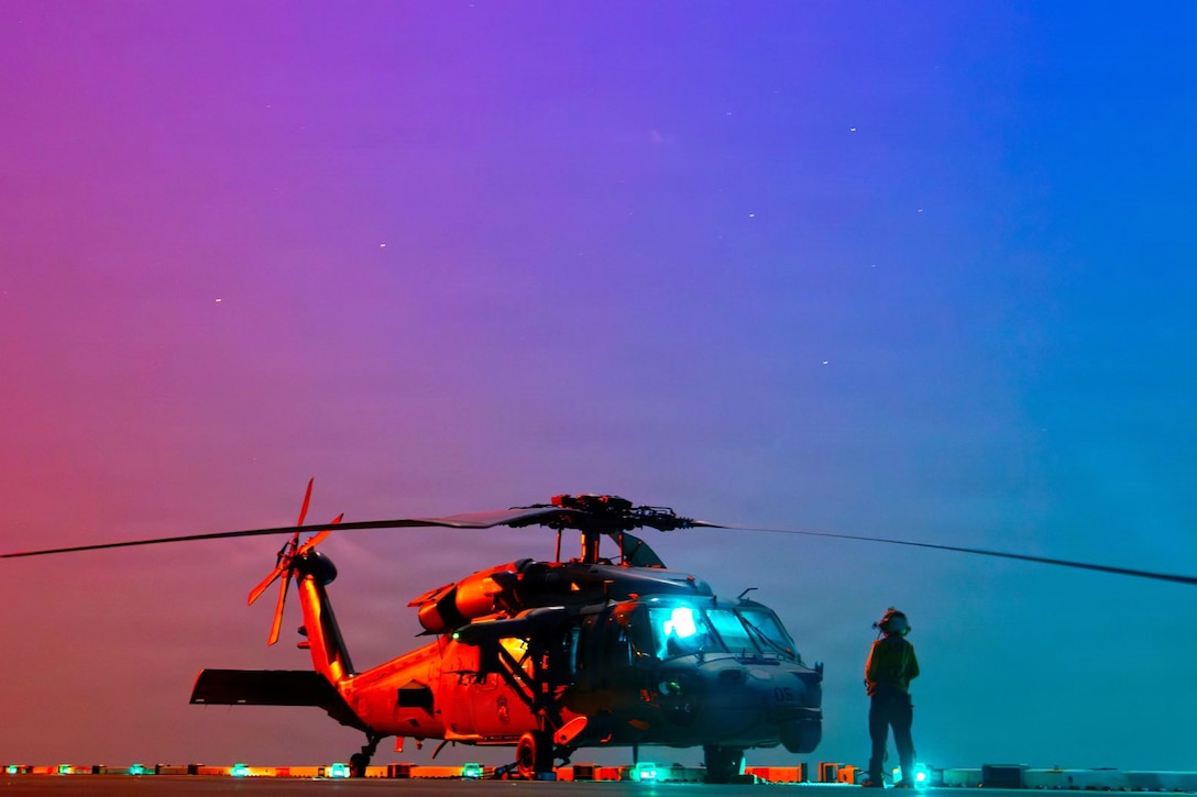 A sailor stands in front of a parked helicopter aboard a ship illuminated by a stary orange, pink and blue sky.
