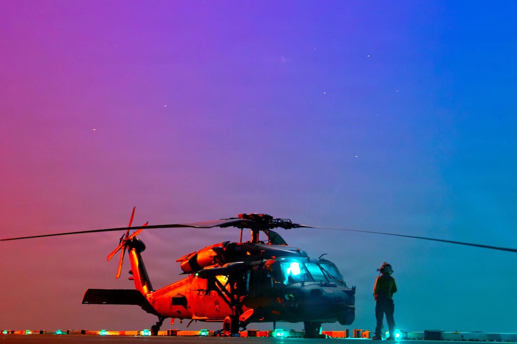 A sailor stands in front of a parked helicopter aboard a ship illuminated by a stary orange, pink and blue sky.