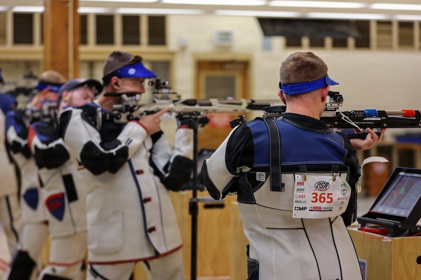 A row of men dressed in competitive rifle shooting gear hold rifles in their hands and stand in the shooting position.