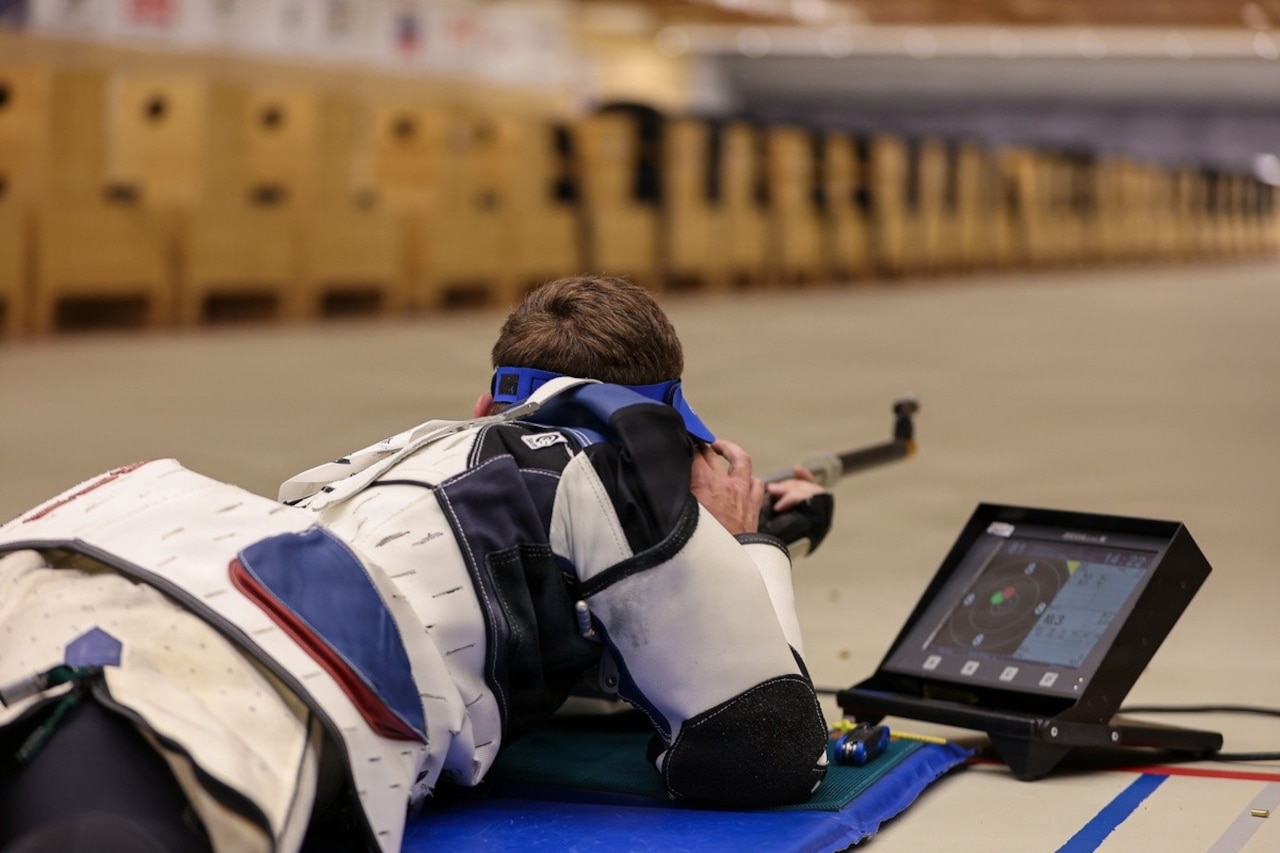 A man dressed in competitive rifle shooting gear is lying down while holding a rifle in the shooting position.