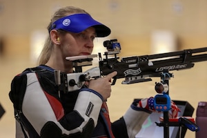 A woman dressed in competitive rifle shooting gear looks at a target and holds a rifle in the shooting position.