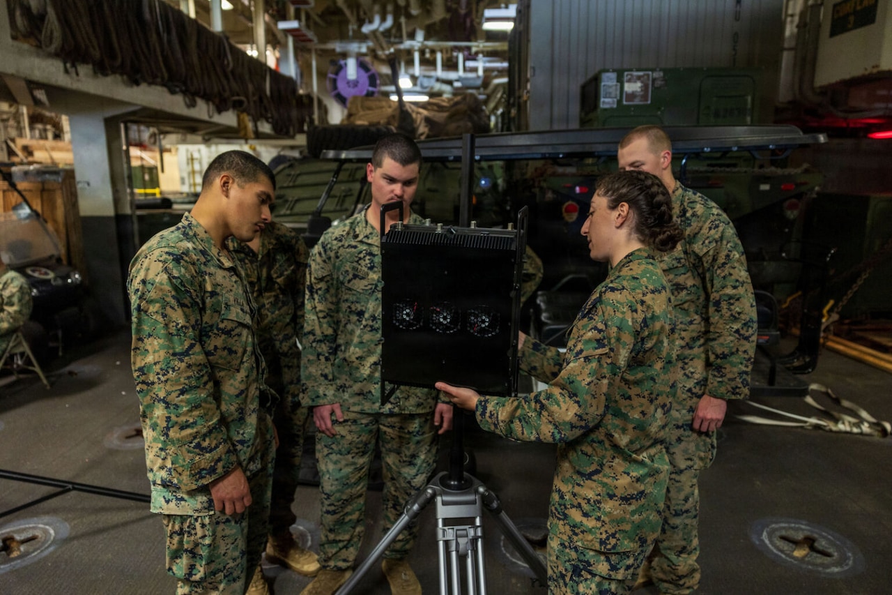 Three men and a woman wearing camouflage military uniforms examine a piece of military equipment while aboard a ship. There is additional military equipment in the background.