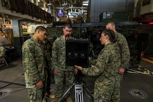 Three men and a woman wearing camouflage military uniforms examine a piece of military equipment while aboard a ship. There is additional military equipment in the background.