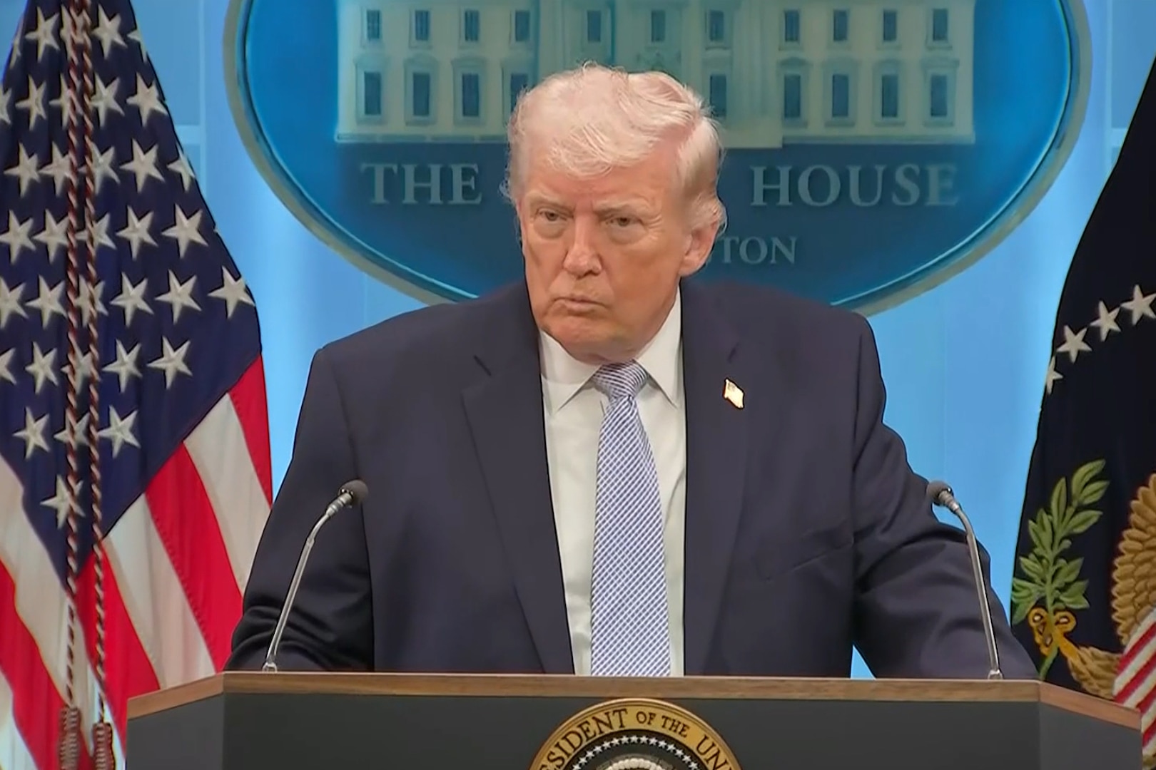A man in a business suit speaks at a lectern, with flags and a White House seal on a blue wall in the background.