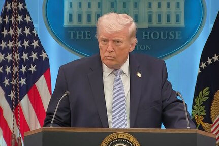 A man in a business suit speaks at a lectern, with flags and a White House seal on a blue wall in the background.