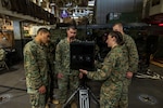 Three men and a woman wearing camouflage military uniforms examine a piece of military equipment while aboard a ship. There is additional military equipment in the background.