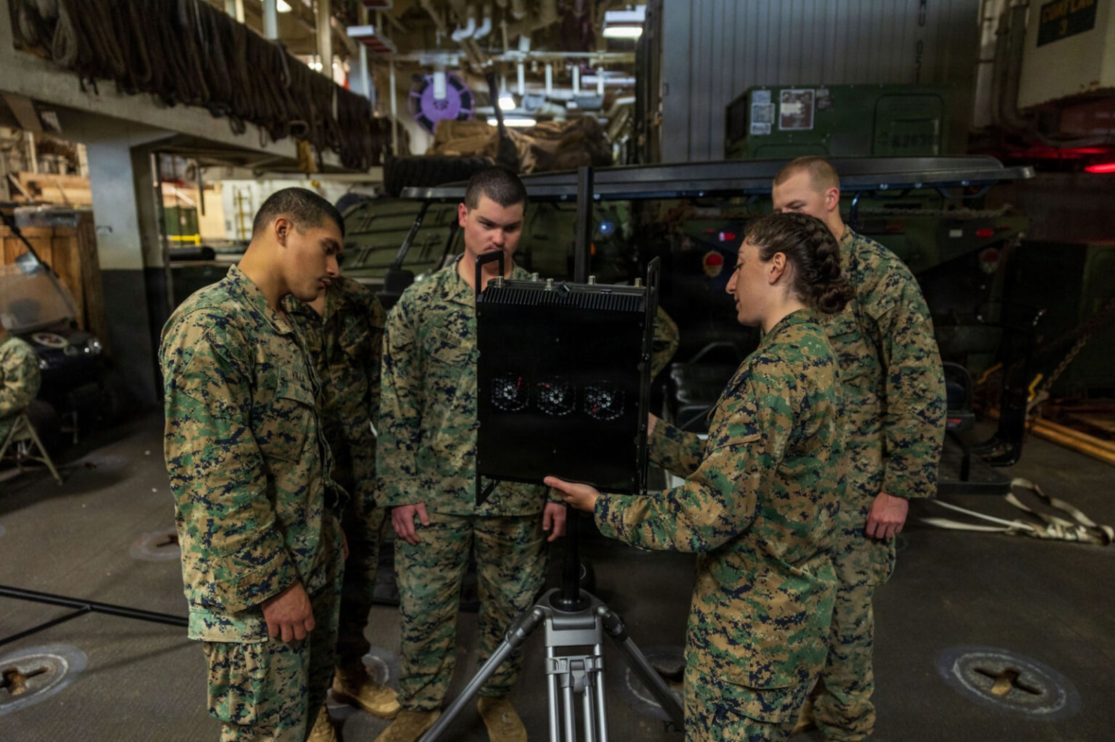 Three men and a woman wearing camouflage military uniforms examine a piece of military equipment while aboard a ship. There is additional military equipment in the background.